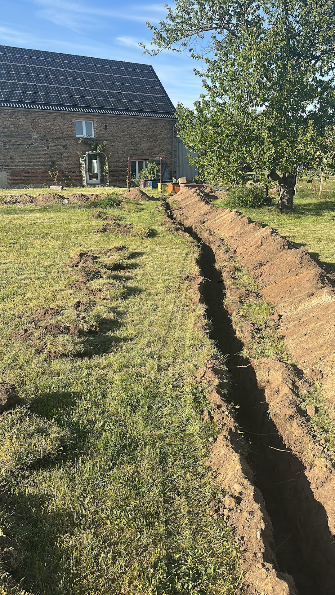 Langer offener Graben im Garten von The Farm für eine neue Wasserleitung, im Hintergrund das Klinkergebäude mit Solardach