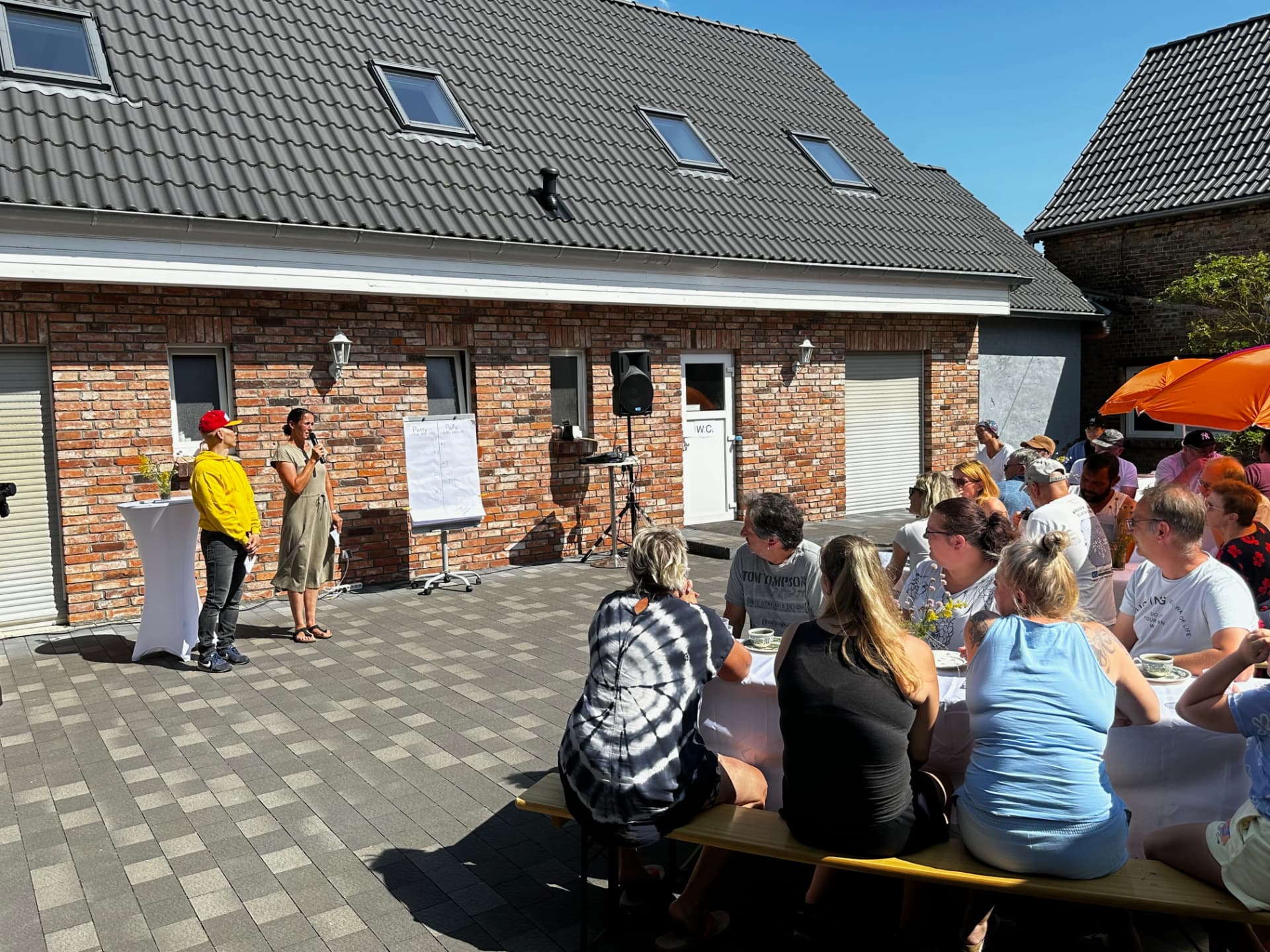 Offene Veranstaltung im Hof: Gäste an langen Tischen, Moderation mit Mikrofon und Flipchart vor der Klinkerwand