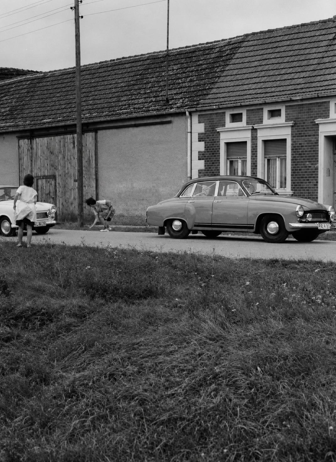 Historische Schwarzweiß-Aufnahme der Dorfstraße in Bräsen mit Wartburg-Limousinen vor Scheune und Klinkerhaus